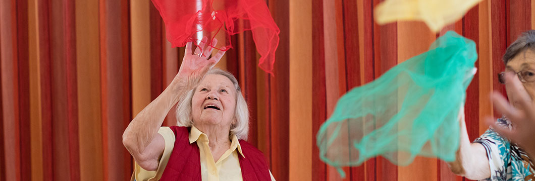 Seine Seniorin führt mit weiteren Senioren Bewegungsübungen mit Tüchern aus.