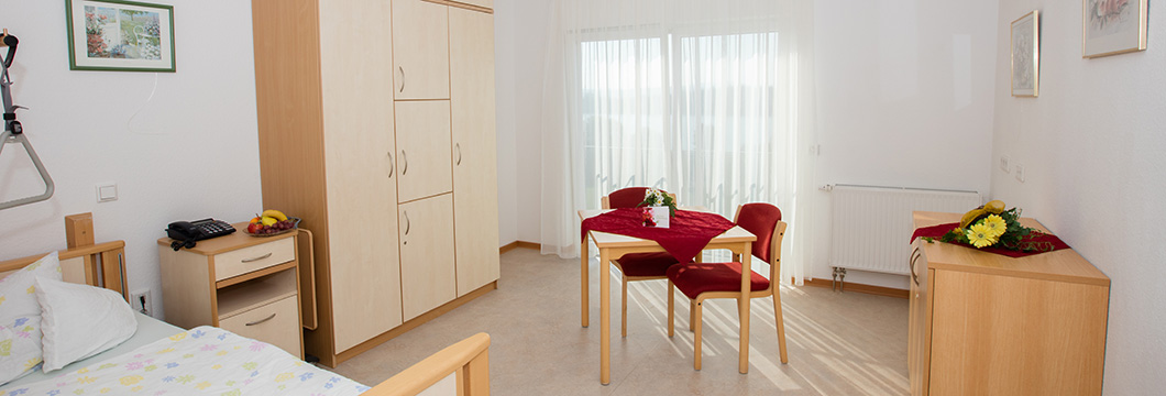Das Zimmer ist geräumig mit einen Pflegebett, einem Tisch mit Stühlen sowie Schränken eingerichtet.