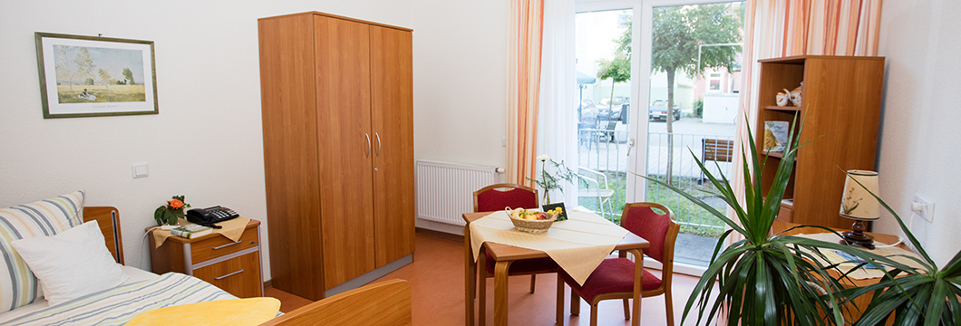 "Dargestellt ist die Einrichtung eines privaten Zimmer im Seniorenheim mit Bett, Tisch, Stühle und Schränken sowie einem großen hellen Fenster."