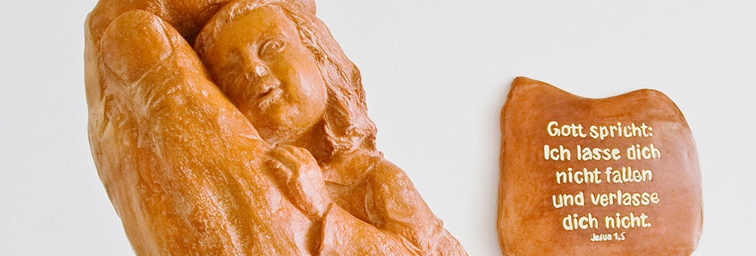 Eine Tonfigur stellt Hände da, in die sich ein Kind lehnt und geborgen fühlt. Dahinter hängt eine Tontafel mit dem Vers aus Josua 1,5: Gott spricht: Ich lasse dich nicht fallen und verlasse dich nicht.